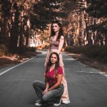 Two women pose confidently on a scenic forest road in casual attire, surrounded by trees.